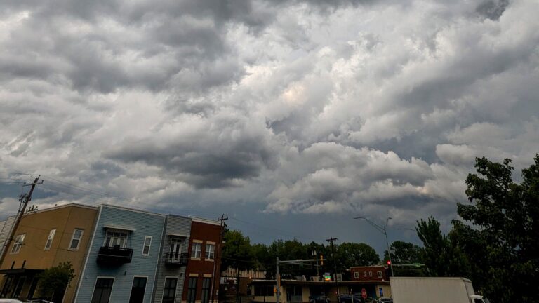 Dark storm clouds over Topeka, Kansas cityscape, with trees and buildings swaying in strong winds, hinting at severe weather and potential tornadoes approaching in the early evening