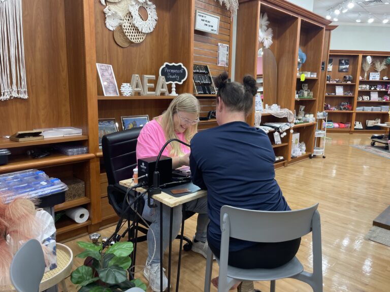 Linked by AEA employee attaching permanent jewelry on a customer during girls' day out event in Topeka. Shelves with local gifts and wellness products in background.