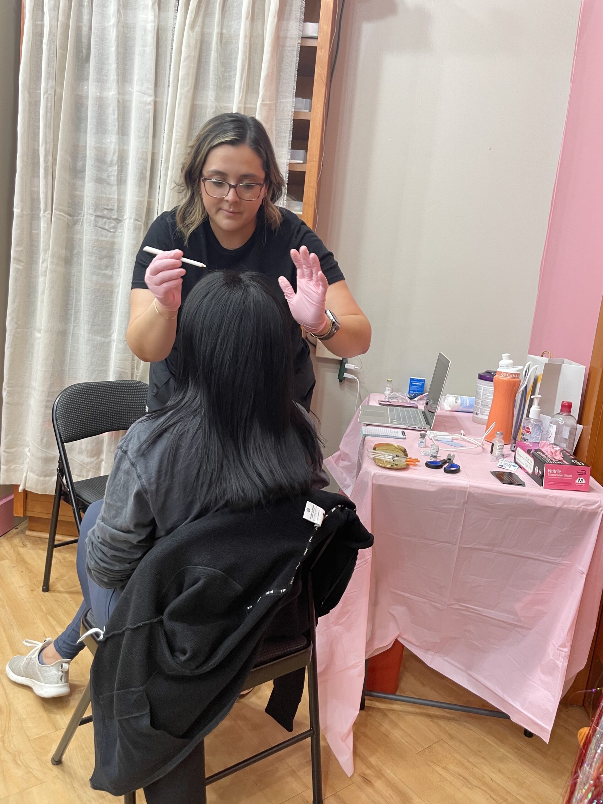 Linked by AEA employee attaching permanent jewelry on a customer during girls' day out event in Topeka. Shelves with local gifts and wellness products in background.