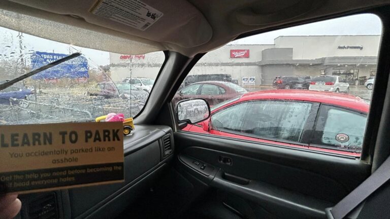 An unofficial parking ticket featuring humorous phrases such as 'You parked like an asshole' and 'You suck at parking,' designed to resemble an official citation. The ticket is placed on the windshield of a car parked awkwardly in a shopping center lot, highlighting poor parking habits.