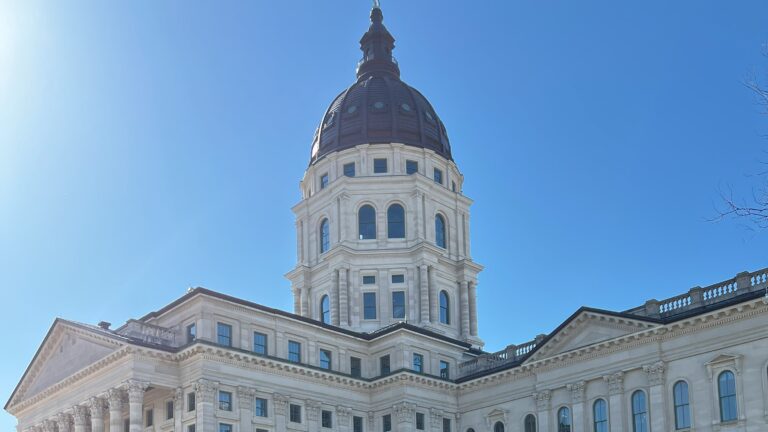 Photograph of the Kansas State Capitol building in Topeka, featuring its iconic green copper dome and classical architecture, set against a vibrant blue sky with scattered white clouds.