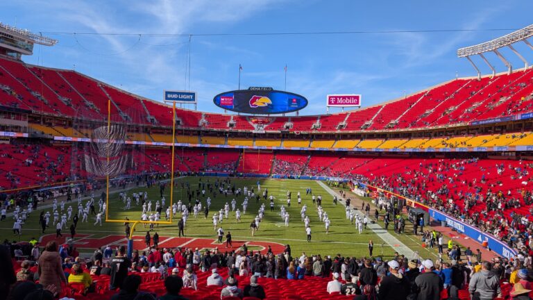 A packed Arrowhead Stadium under the blue sky, filled with roaring fans wearing a mix of crimson and blue for the Kansas Jayhawks and black and gold for the Colorado Buffaloes. The bright stadium lights illuminate the field as KU players celebrate a touchdown, while CU defenders regroup. The scoreboard in the background displays KU's lead, capturing the energy and intensity of the Big 12 showdown.