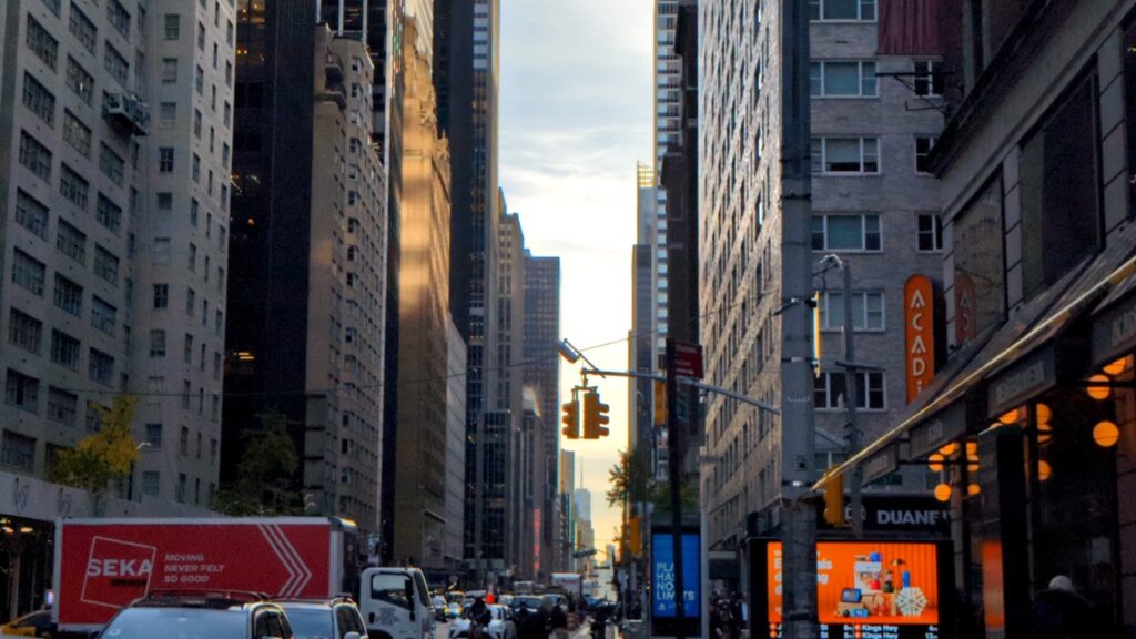 A bustling New York City street during the day, lined with iconic skyscrapers and classic brownstone buildings. Yellow taxis weave through traffic, while pedestrians cross the street and stroll along busy sidewalks. The scene captures the dynamic energy and charm of Manhattan in full daylight