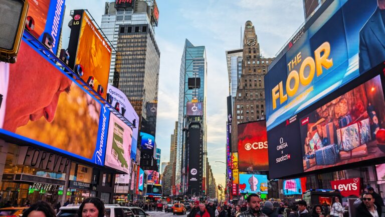 A vibrant view of Times Square at night, illuminated by towering digital billboards and neon signs. Crowds of people fill the streets, creating a lively and energetic atmosphere. Iconic advertisements and yellow taxis are visible, capturing the heart of New York City's hustle and bustle