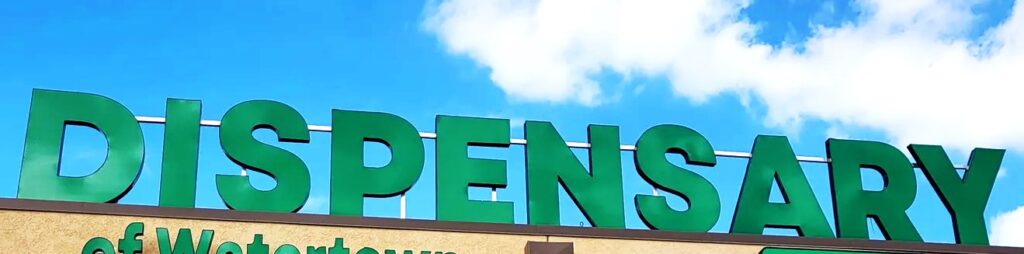 Close-up of a large, green 'Dispensary' sign mounted on the exterior of a building against a bright blue sky with scattered white clouds. The bottom part of the sign includes additional text partially visible, identifying the location