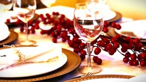 A festive holiday table setting featuring a clear wine glass in focus. The table is decorated with red berry garlands, golden charger plates, white dinner plates, and neatly folded white napkins tied with twine for a rustic touch. The scene evokes a warm and elegant Christmas dining atmosphere.