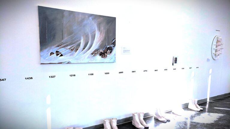 Gallery wall displaying contemporary art in Topeka, Kansas, featuring a dramatic wave painting, numbered timeline installation, and sculptural white boots as part of a thought-provoking exhibit