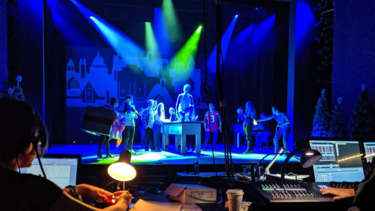 Behind-the-scenes view of a live theater production in Topeka, Kansas, featuring performers on stage under colorful lights with stage crew operating sound and lighting equipment in the foreground.