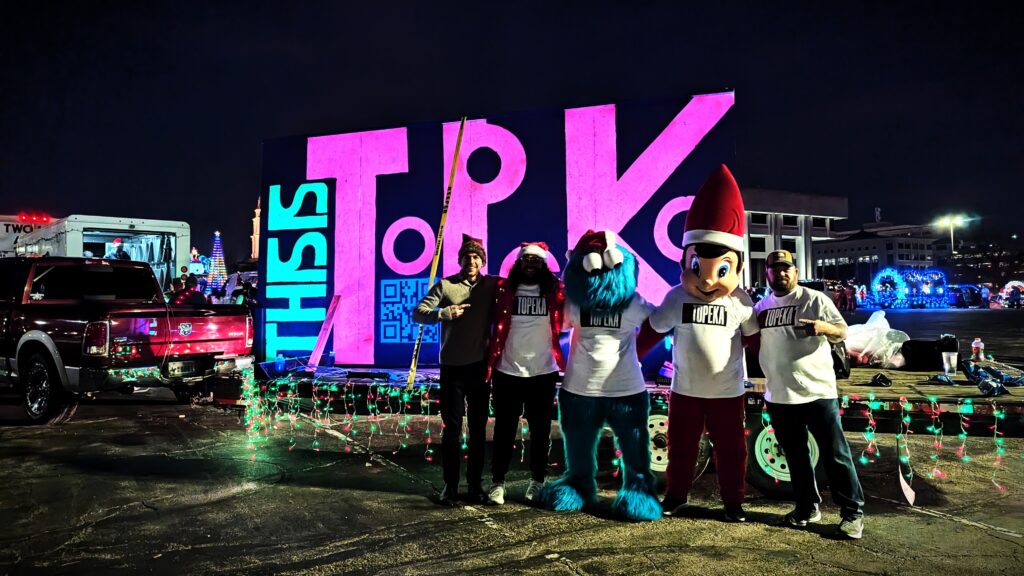 A group of people posing in front of a colorful 'This Is Topeka' float during the Topeka Christmas Light Parade. From left to right: artist Mike G, Tevyn Harmon, Cookie Monster, Elf on the Shelf, and Jeff Fogg. The float features vibrant lights and large block letters spelling 'Topeka.' All are wearing shirts with 'Topeka' printed on them, with some in festive costumes. A decorated red RAM truck and additional holiday floats are visible in the background