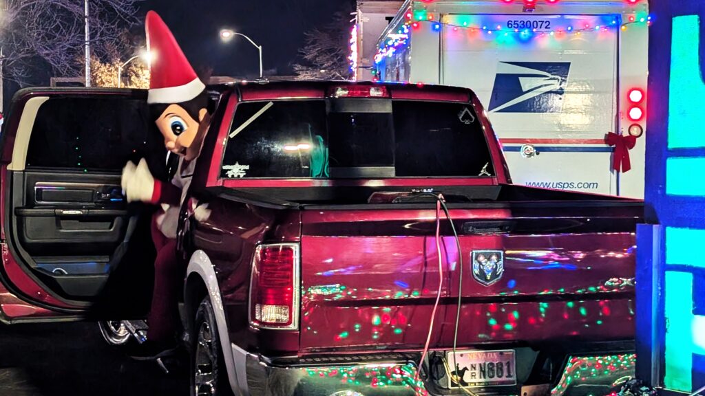 Festive Elf on the Shelf character stepping out of a red RAM 1500 truck adorned with Christmas lights during the Topeka Christmas Light Parade, with a decorated USPS truck visible in the background