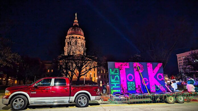 Festive holiday float from ThisIsTopeka.com lit up with neon letters and Christmas lights, towed by a red RAM 1500 truck in front of the illuminated Kansas State Capitol at night during the Topeka Christmas Light Parade