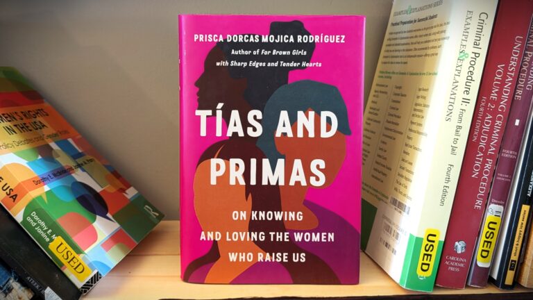 Photograph of the book Tías and Primas: On Knowing and Loving the Women Who Raise Us by Prisca Dorcas Mojica Rodríguez. The book cover features silhouettes of women in bold, earthy colors layered over a vivid pink background. To the right, the book rests on a shelf next to textbooks on women's rights, criminal procedure, and other subjects, emphasizing a context of education and empowerment.