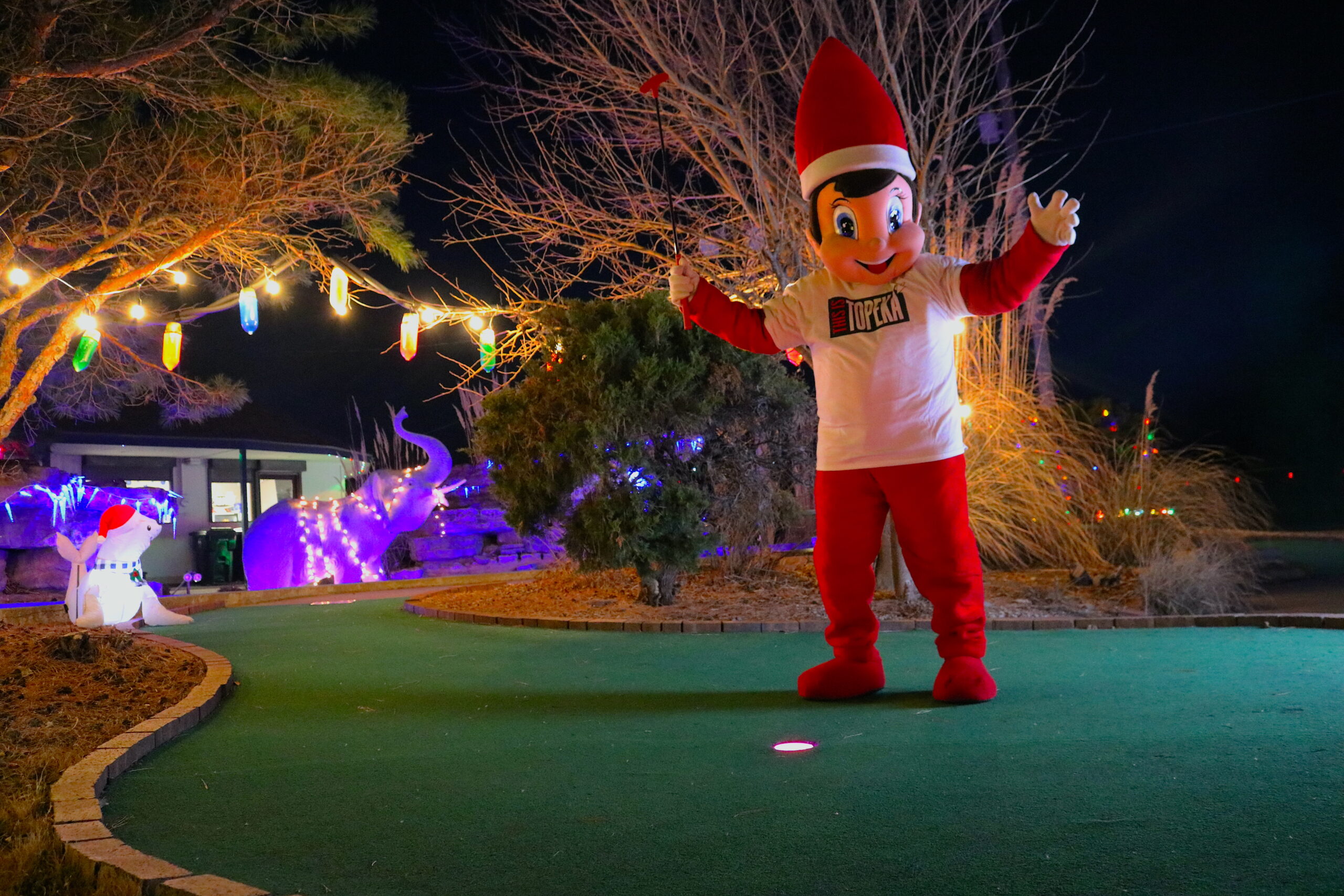 Corner 21 Sports Santa's Village featuring glow-in-the-dark golf balls, vibrant Christmas lights at a mini golf hole, and Elfie the Elf joyfully holding a golf club under festive decorations