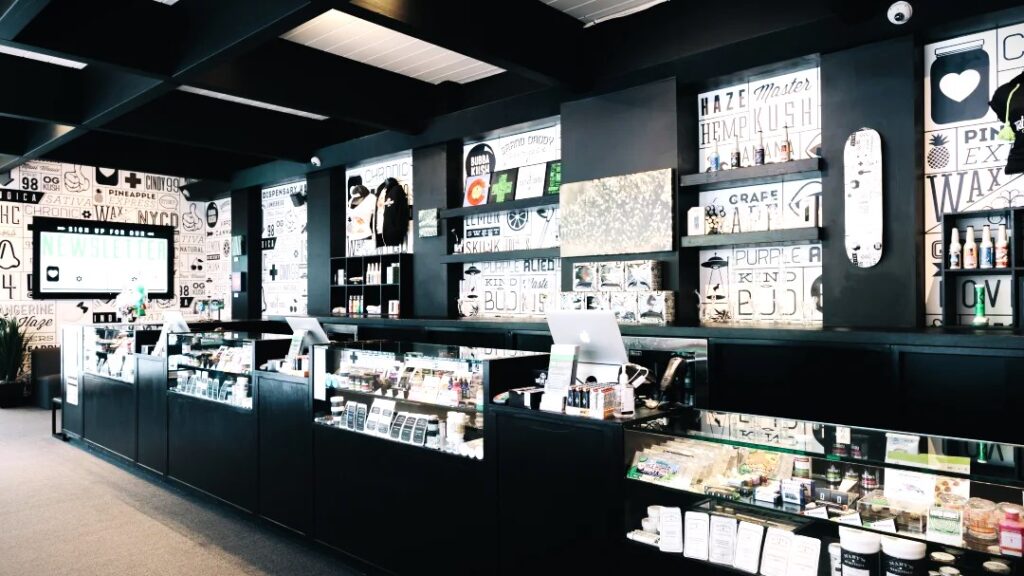 The interior of a modern cannabis dispensary featuring sleek black counters with glass display cases showcasing various cannabis products. The walls are decorated with bold typography and graphics related to cannabis strains and products, creating a stylish and inviting atmosphere.