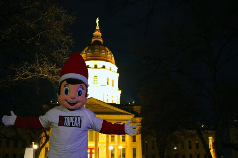 Elfie the Christmas elf standing in front of the State Capitol building, spreading holiday cheer with festive lights in the background