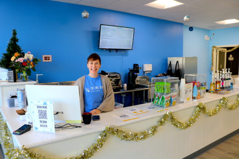 Kayla, owner of To The Stars Play Cafe, smiling at the front desk with a welcoming cafe behind her, featuring coffee, snacks, and a vibrant play area for kids.